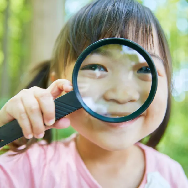 虫眼鏡を持って笑顔でこちらを見る子ども。子どもの不調の背景や原因を深く観察するイメージ